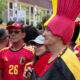 Group of Belgian football fans in red and yellow, with a woman in a tall pink-and-yellow hat in the foreground at a street event.