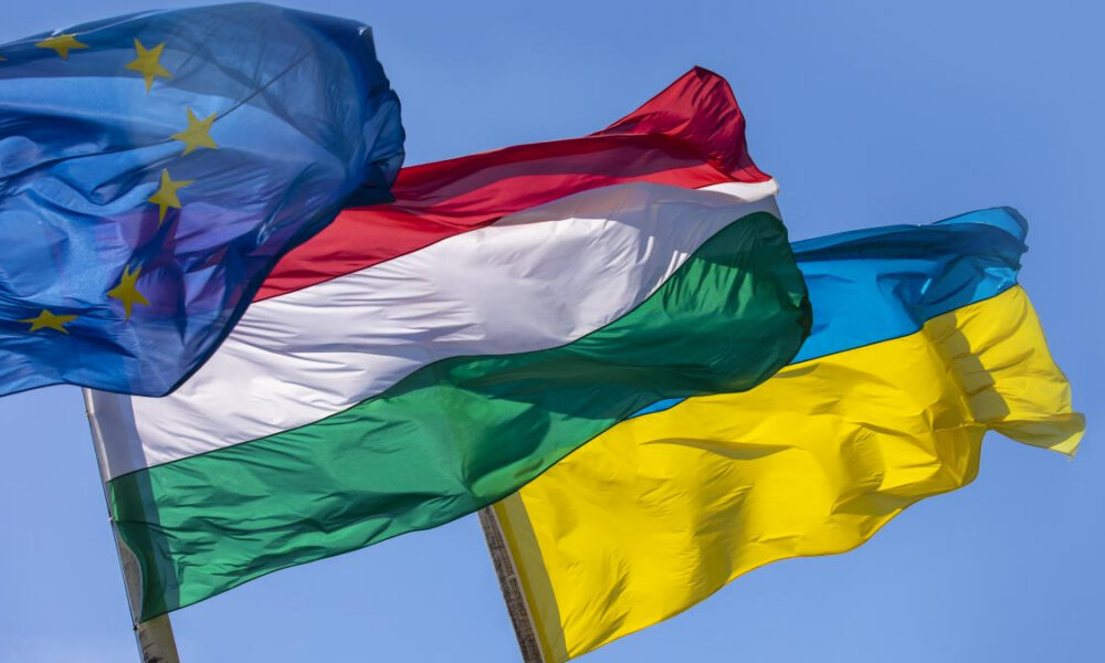 EU flag with yellow stars, Hungary flag, and Ukraine flag fluttering in a clear blue sky.
