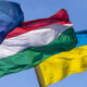 EU flag with yellow stars, Hungary flag, and Ukraine flag fluttering in a clear blue sky.