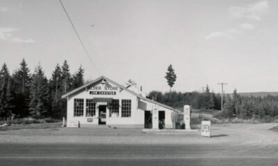 Vintage gas station/store set along a dirt road, surrounded by evergreen trees; pumps and an open sign outside in black-and-white photo.”,