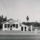 Vintage gas station/store set along a dirt road, surrounded by evergreen trees; pumps and an open sign outside in black-and-white photo.”,
