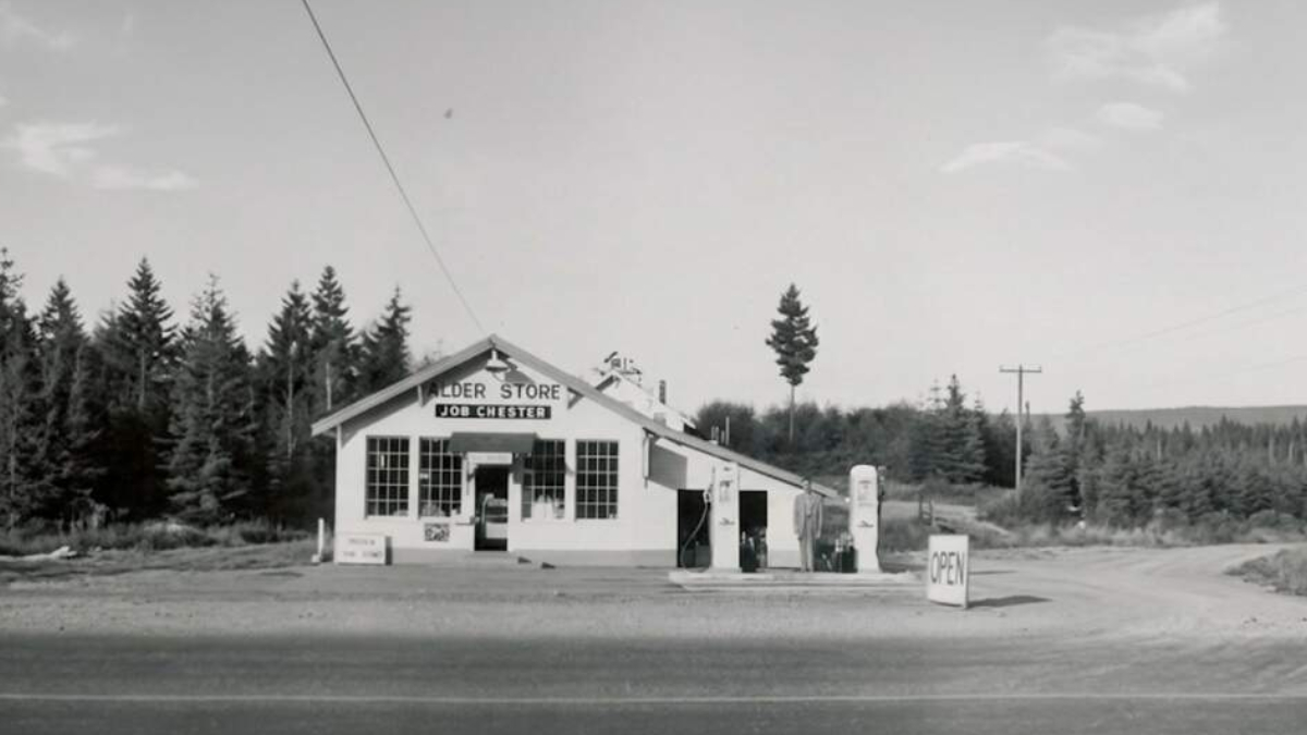 Vintage gas station/store set along a dirt road, surrounded by evergreen trees; pumps and an open sign outside in black-and-white photo.”,