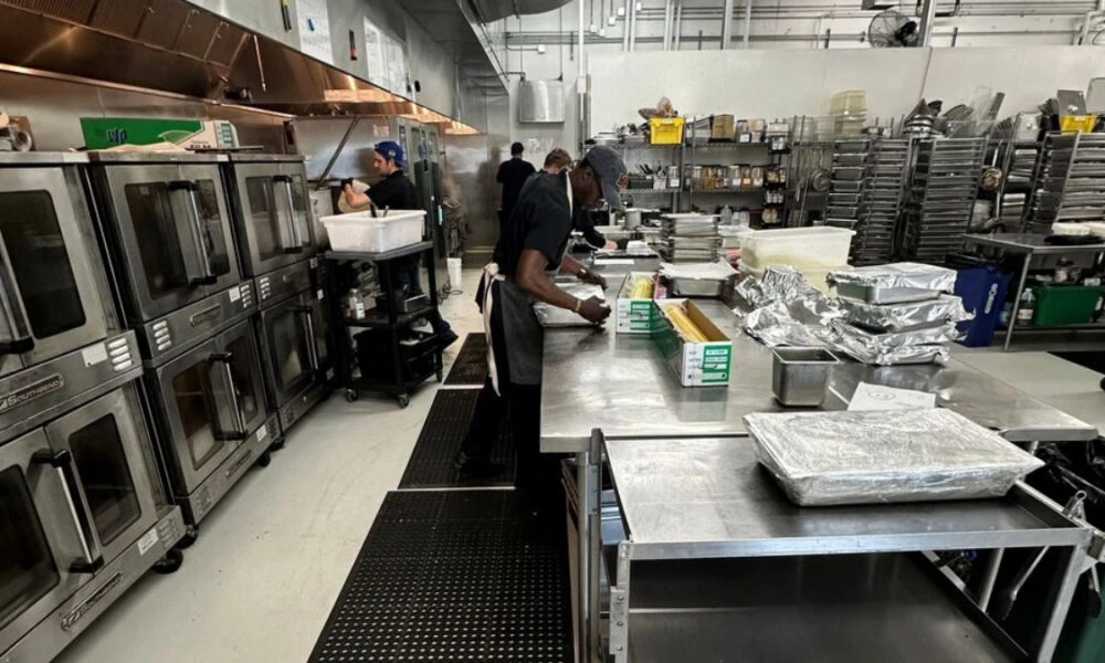 Commercial kitchen scene: stainless prep tables with staff preparing food, large industrial ovens on the left.