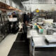 Commercial kitchen scene: stainless prep tables with staff preparing food, large industrial ovens on the left.