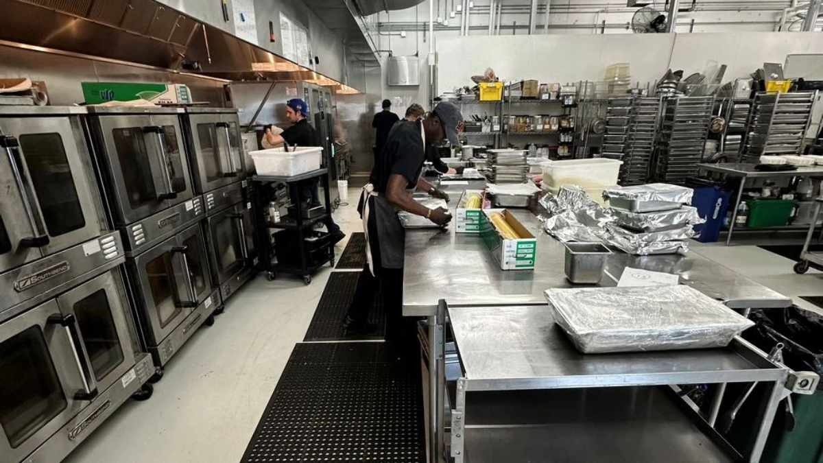 Commercial kitchen scene: stainless prep tables with staff preparing food, large industrial ovens on the left.