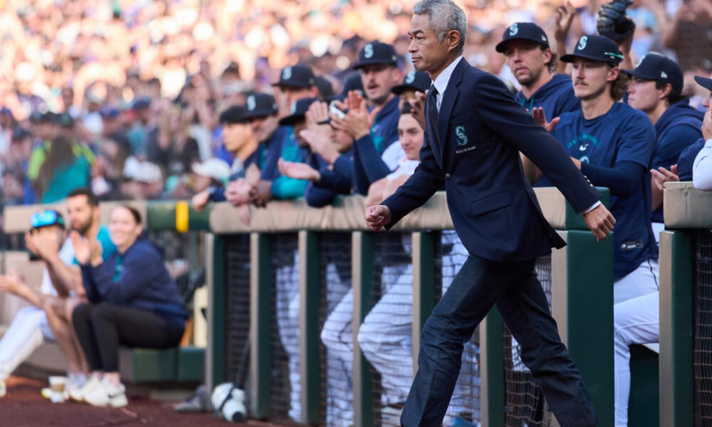 Former president Barack Obama in a navy blazer walking along the dugout fence as players cheer in a baseball stadium at a game.