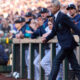 Former president Barack Obama in a navy blazer walking along the dugout fence as players cheer in a baseball stadium at a game.