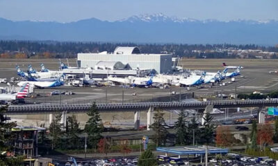 Wide view of an airport with Alaska Airlines planes at gates, a nearby elevated highway, and distant snow-capped mountains.