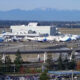 Wide view of an airport with Alaska Airlines planes at gates, a nearby elevated highway, and distant snow-capped mountains.