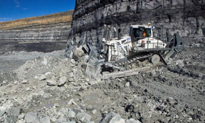 Large industrial rock-cutting machine operating in an open-pit quarry with piles of rubble and a cliff face nearby, blue sky above.