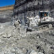 Large industrial rock-cutting machine operating in an open-pit quarry with piles of rubble and a cliff face nearby, blue sky above.