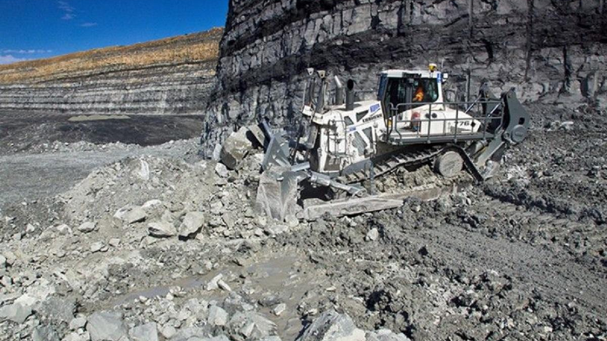 Large industrial rock-cutting machine operating in an open-pit quarry with piles of rubble and a cliff face nearby, blue sky above.