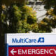 MultiCare sign with a blue medical cross and a red EMERGENCY banner pointing left, outdoors.