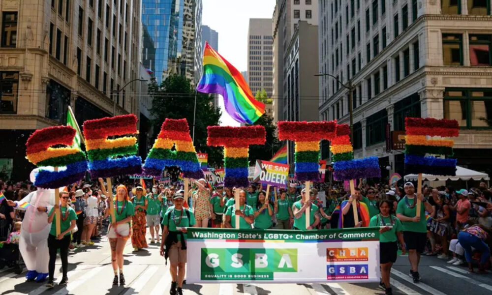 Rainbow letters spelling EQUALITY carried above a Pride march in a city street, with supporters holding signs and a banner for LGBTQ+ chamber of commerce.