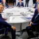 Two men in suits sit at a round conference table during a formal international meeting.