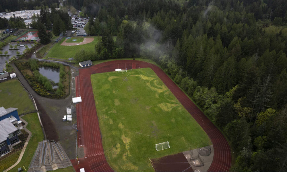 Aerial view of a red running track encircling a green sports field, with goalposts and nets nearby and dense trees in the background.