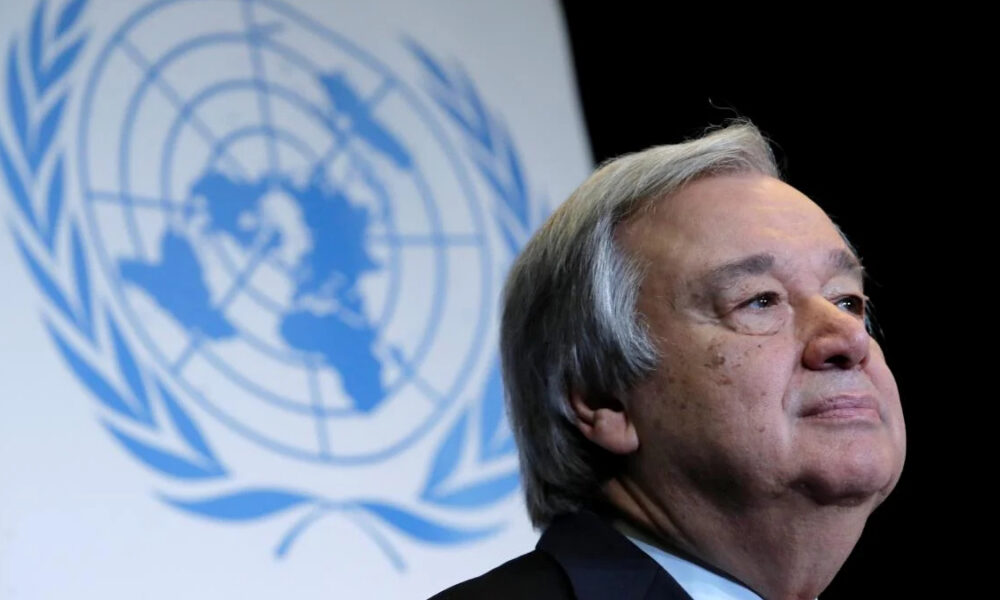 Older man in a suit standing at a UN event with the United Nations emblem in blue behind him.