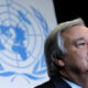 Older man in a suit standing at a UN event with the United Nations emblem in blue behind him.