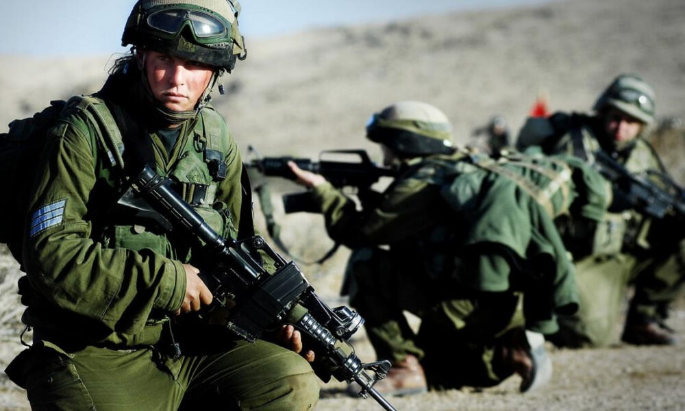 Soldier in green combat gear crouching with rifle in a desert training exercise, others kneeling in the background with rifles ready.