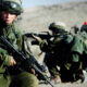 Soldier in green combat gear crouching with rifle in a desert training exercise, others kneeling in the background with rifles ready.