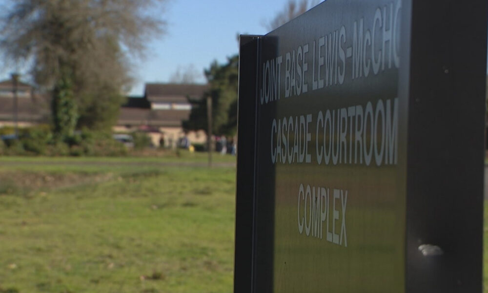 Close-up of a dark outdoor sign reading Cascade Courtroom Complex, with grass and trees and blurred campus buildings in the background.