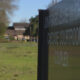 Close-up of a dark outdoor sign reading Cascade Courtroom Complex, with grass and trees and blurred campus buildings in the background.