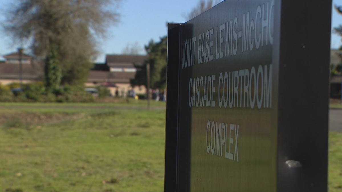 Close-up of a dark outdoor sign reading Cascade Courtroom Complex, with grass and trees and blurred campus buildings in the background.