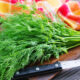 Fresh dill bouquet tied with string on a wooden cutting board beside a kitchen knife and a red-yellow checkered towel.
