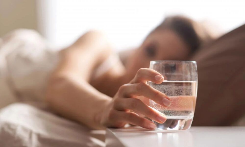 Person lying in bed reaching for a glass of water on a bedside table.