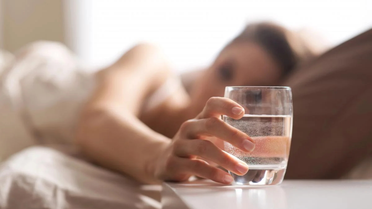Person lying in bed reaching for a glass of water on a bedside table.