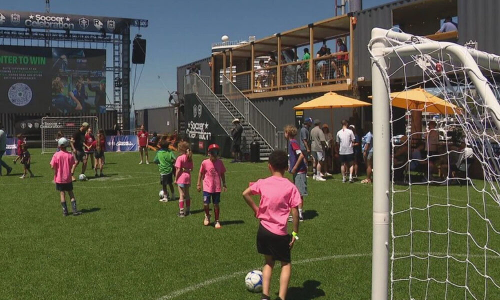 Children in pink shirts play soccer on a sunny field with a small goal and a crowd watching from a balcony area and orange umbrellas nearby, event atmosphere evident.
