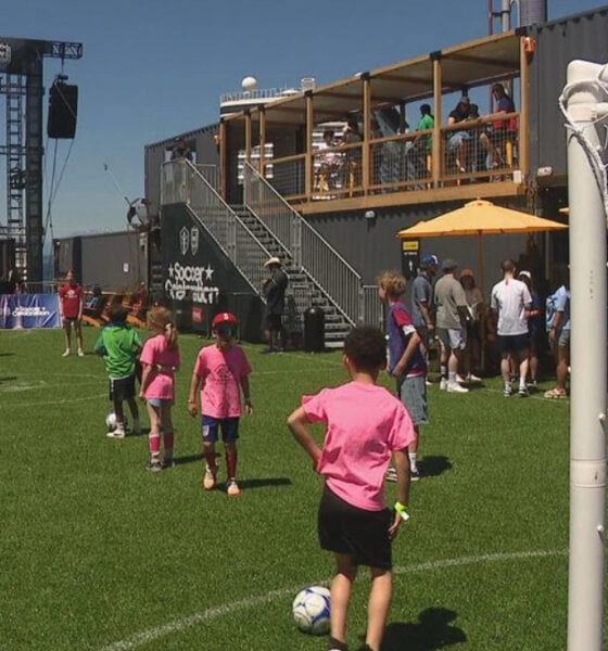 Children in pink shirts play soccer on a sunny field with a small goal and a crowd watching from a balcony area and orange umbrellas nearby, event atmosphere evident.