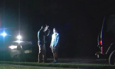 Two men stand and talk beside a car with flashing police lights at night. A second vehicle is parked nearby.
