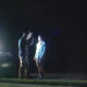 Two men stand and talk beside a car with flashing police lights at night. A second vehicle is parked nearby.