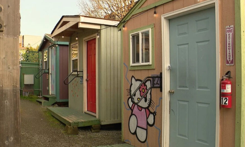 Row of colorful small huts along a gravel alley; beige wall features a Hello Kitty mural beside a blue door and red door.
