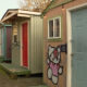 Row of colorful small huts along a gravel alley; beige wall features a Hello Kitty mural beside a blue door and red door.