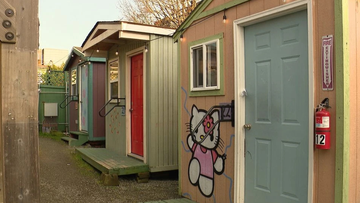 Row of colorful small huts along a gravel alley; beige wall features a Hello Kitty mural beside a blue door and red door.