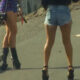 Two women walk away on a paved road; one wears black high-heeled boots and a purple patterned outfit, the other in denim shorts and black heeled sandals.
