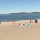 Busy sunny beach with people sunbathing on towels near the blue ocean. Boats visible in the distance.