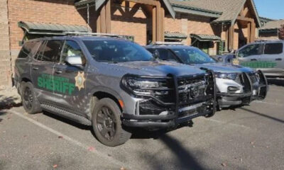 Two sheriff patrol SUVs with front-end damage parked in a lot outside brick buildings.