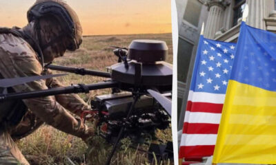 Soldier in camouflage crouches beside a mounted weapon in a grassy field at sunset, with US and Ukrainian flags nearby.