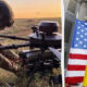 Soldier in camouflage crouches beside a mounted weapon in a grassy field at sunset, with US and Ukrainian flags nearby.