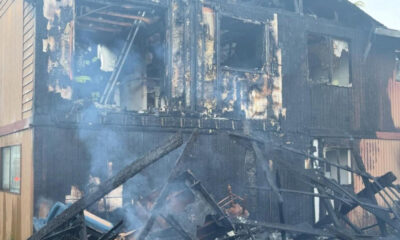 Charred two-story building with extensive fire damage and smoke, firefighters visible inside the wreckage