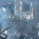 Charred two-story building with extensive fire damage and smoke, firefighters visible inside the wreckage