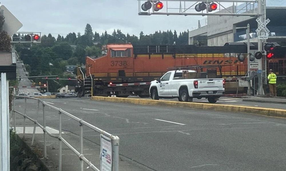 Orange BNSF locomotive crossing a street, with a white pickup truck in front and a worker in a high-visibility vest nearby.