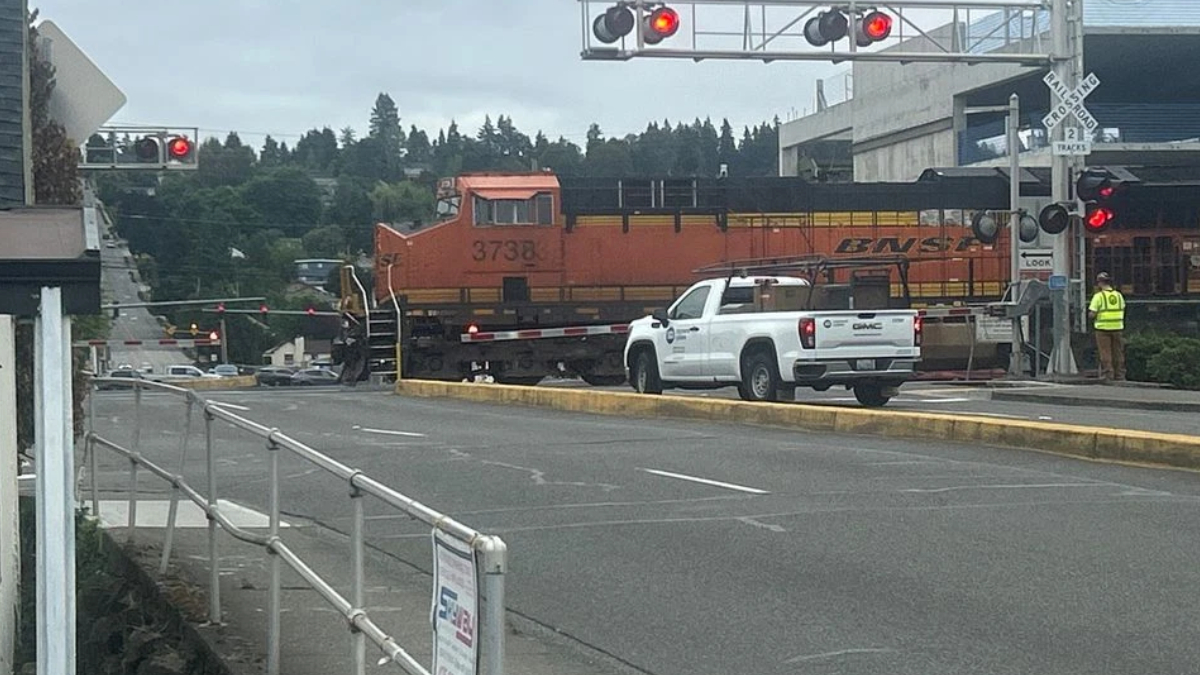 Orange BNSF locomotive crossing a street, with a white pickup truck in front and a worker in a high-visibility vest nearby.