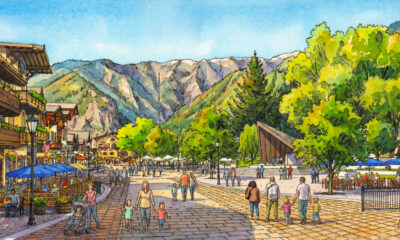 Bustling mountain village square with outdoor cafes, pedestrians, and families against a backdrop of green hills and distant mountains.