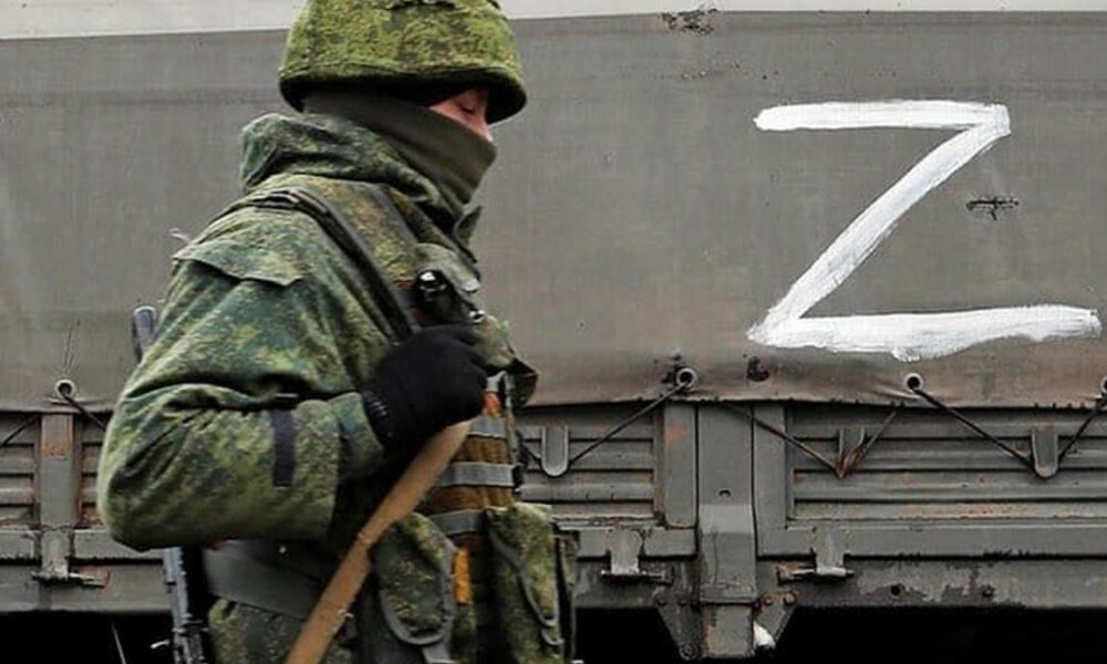 Camouflaged soldier with face covered stands beside a gray train car painted with a large white Z.