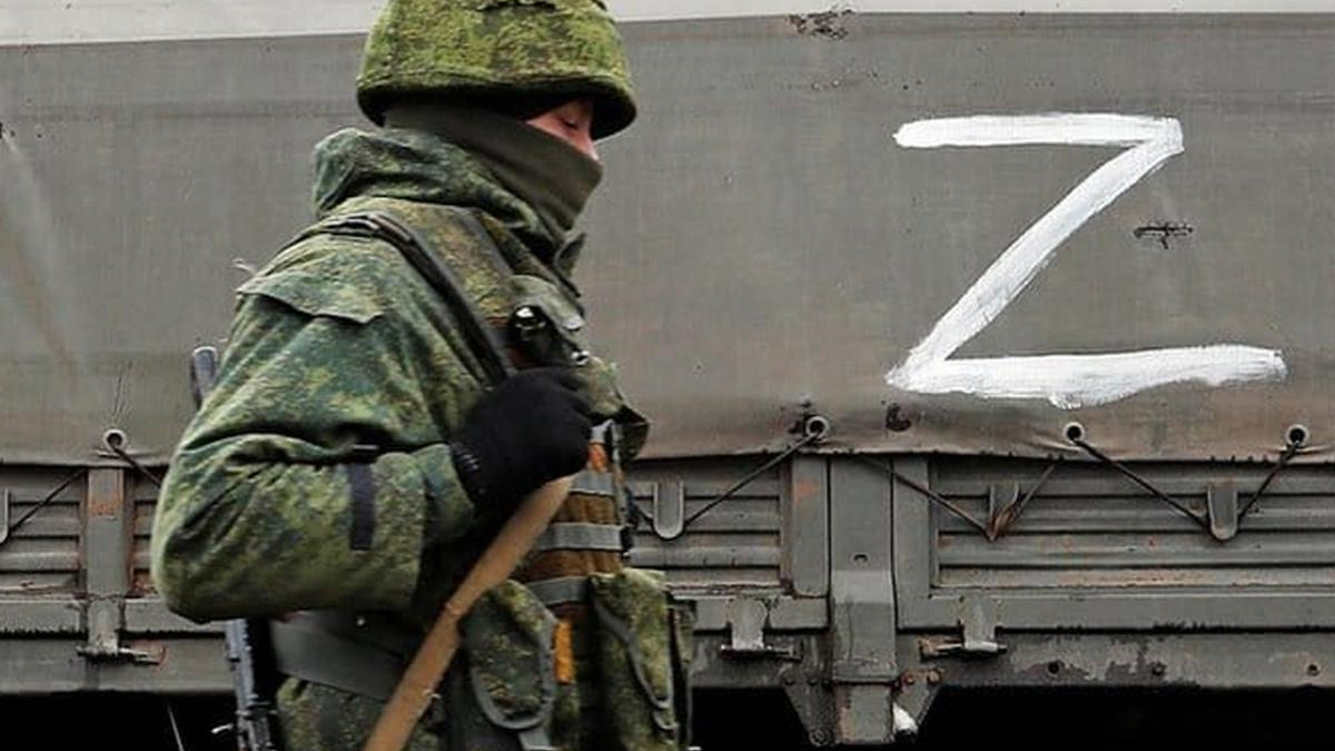 Camouflaged soldier with face covered stands beside a gray train car painted with a large white Z.