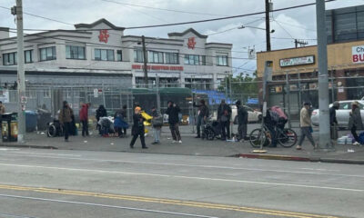 People line up along a sidewalk beside a chain-link fence in front of a building, with bags and belongings scattered nearby.